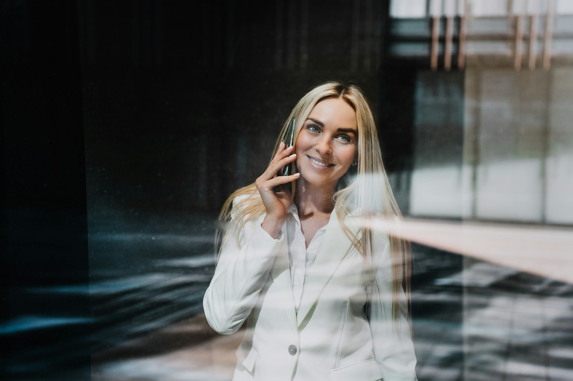 Beautiful blonde businesswoman in suite talks by phone, broad smiles, looks up through the window