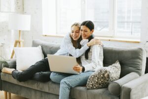 two young female students Asian and blonde sitting on couch and looking at laptop, watching movie or