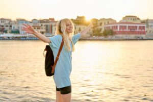 Happy blonde female enjoying in headphones with backpack on sea promenade